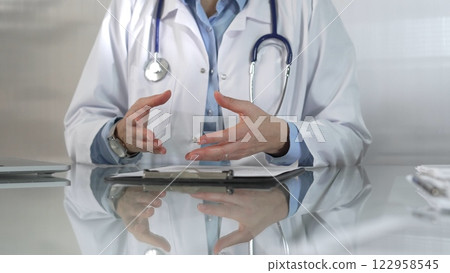Doctor woman wearing a blue blouse, stethoscope and lab coat is gesturing with hands while explaining a diagnosis to a patient across a glass desk with a laptop. Medicine concept Doctor woman wearing a blue blouse, stethoscope and lab coat is gesturing with hands while explaining a diagnosis to a patient across a glass desk with a laptop. Medicine concept 122958545