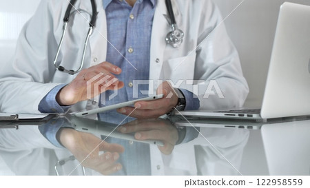 Doctor man, wearing a blue shirt under a medical white coat, is using digital tablet in modern healthcare setting, interacting with patient data, close-up. Medicine and health care Doctor man, wearing a blue shirt under a medical white coat, is using digital tablet in modern healthcare setting, interacting with patient data, close-up. Medicine and health care 122958559