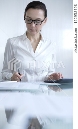 Businesswoman or accountant, wearing white casual blouse and glasses, is working on tax calculations, note taking, and financial reports in the office. Audit and taxes in business Businesswoman or accountant, wearing white casual blouse and glasses, is working on tax calculations, note taking, and financial reports in the office. Audit and taxes in business 122958598