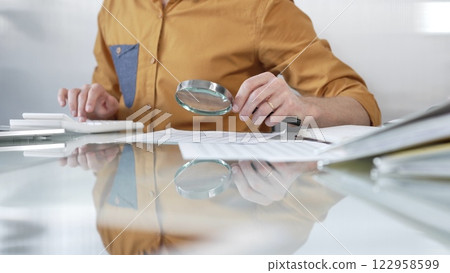 Businessman accountant wearing casual yellow shirt carefully reviewing financial reports with magnifying glass and calculator, conducting audit and analyzing data in office. Taxes, audit in business Businessman accountant wearing casual yellow shirt carefully reviewing financial reports with magnifying glass and calculator, conducting audit and analyzing data in office. Taxes, audit in business 122958599