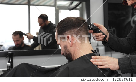 Skilled barber trimming client's hair using electric razor, creating contemporary hairstyle in modern barbershop setting with precise, professional technique 122958611