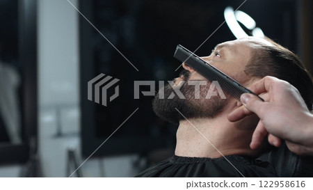 Skilled barber carefully combing thick beard of male client, providing professional grooming service in traditional barbershop setting with precision and attention to detail Skilled barber carefully combing thick beard of male client, providing professional grooming service in traditional barbershop setting with precision and attention to detail 122958616