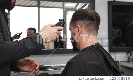 Skilled barber trimming client's hair using electric razor, creating contemporary hairstyle in modern barbershop setting with precise, professional technique 122958619