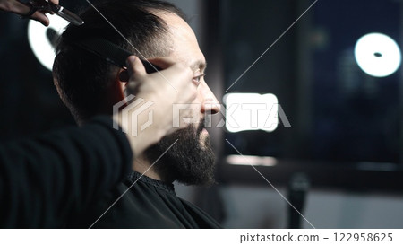 Professional barber trimming client's hair using professional scissors, creating stylish fade for bearded man in modern barbershop setting 122958625