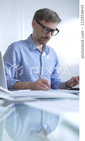 Businessman or accountant, wearing blue casual shirt and glasses, is working on tax calculations, note taking, and financial reports in the office. Audit and taxes in business Businessman or accountant, wearing blue casual shirt and glasses, is working on tax calculations, note taking, and financial reports in the office. Audit and taxes in business 122958645