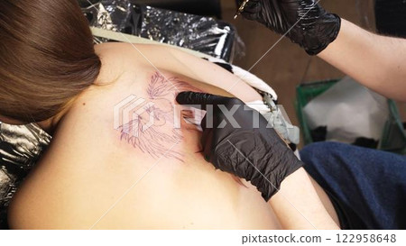 Professional tattoo artist wearing black gloves drawing intricate design on woman's back while working in sleek, well lit tattoo studio with modern equipment 122958648