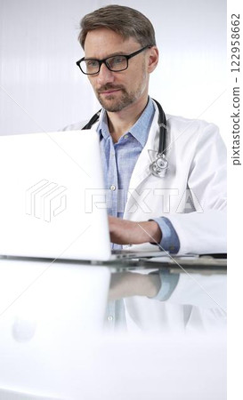 Doctor man in a blue shirt under a medical coat is using technology in medicine. Portrait of physician typing on a laptop on a glass desk in clinic, vertical view. Medicine and science Doctor man in a blue shirt under a medical coat is using technology in medicine. Portrait of physician typing on a laptop on a glass desk in clinic, vertical view. Medicine and science 122958662