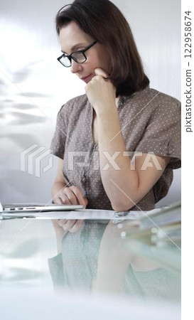 Focused businesswoman in brawn dotted blouse is using laptop and thinking with hand on chin, working on a project in a bright office. Business people concept Focused businesswoman in brawn dotted blouse is using laptop and thinking with hand on chin, working on a project in a bright office. Business people concept 122958674