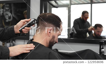 Skilled barber trimming client's hair using electric razor, creating contemporary hairstyle in modern barbershop setting with precise, professional technique Skilled barber trimming client's hair using electric razor, creating contemporary hairstyle in modern barbershop setting with precise, professional technique 122958696