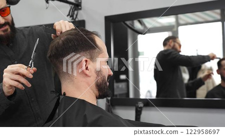 Skilled barber working on male client's hairstyle, carefully combing and preparing precision cut in contemporary barbershop setting Skilled barber working on male client's hairstyle, carefully combing and preparing precision cut in contemporary barbershop setting 122958697