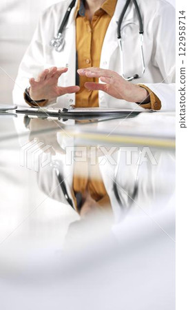 Unknown male doctor wearing a stethoscope, yellow shirt under a lab coat is gesturing with hands overs a glass desk with a laptop in clinic office. Medicine concept 122958714