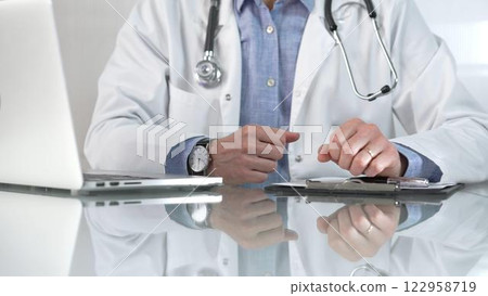 Male doctor wearing a blue shirt, stethoscope and lab coat is gesturing with hands while explaining a diagnosis to a patient across a glass desk with a laptop. Medicine concept 122958719