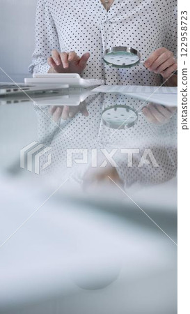 Businesswoman in white dotted blouse is carefully reviewing financial reports with magnifying glass and calculator, conducting audit and analyzing data, vertical view Taxes, audit in business 122958723