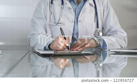 Woman doctor, wearing a blue blouse under a white medical coat, is using a clipboard and taking notes on glass desk in a medical office, close-up. Medicine 122958727