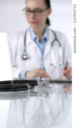 Stethoscope and medication history records form are lying on a glass desk before a doctor working with laptop in clinical office background, vertical close-up view. Medicine and science Stethoscope and medication history records form are lying on a glass desk before a doctor working with laptop in clinical office background, vertical close-up view. Medicine and science 122958732