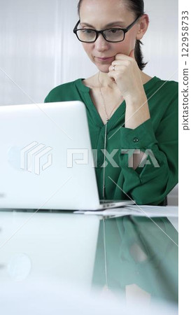 Focused businesswoman in green blouse is using laptop and thinking with hand on chin, working on a project in a bright office, vertical portrait. Business people concept 122958733
