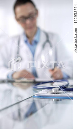 Blue stethoscope and medication history records form are lying on a glass desk before a doctor working with laptop in clinical office background, vertical close-up view. Medicine and science Blue stethoscope and medication history records form are lying on a glass desk before a doctor working with laptop in clinical office background, vertical close-up view. Medicine and science 122958734