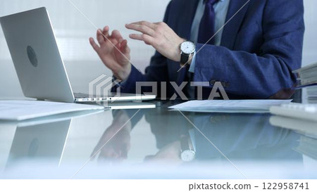 Businessman in blue suit is gesturing with his hands near laptop screen at glass desk while sitting in bright office, online communication, close-up view. Business concept 122958741