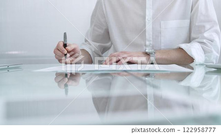 Businessman in white casual shirt is signing a contract at a sleek glass desk in a modern office, showcasing professionalism and focus in his work environment. Business people concept 122958777