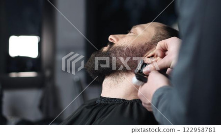 Professional barber trimming client's beard with electric razor, delivering precise styling within contemporary grooming salon setting 122958781