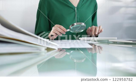 Businesswoman in green blouse carefully reviewing financial reports with magnifying glass and calculator, conducting audit and analyzing data in office. Taxes, audit in business 122958782