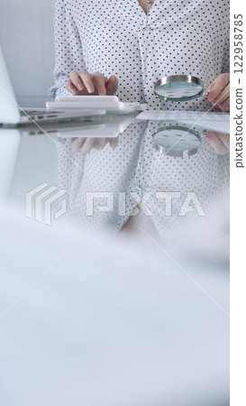 Businesswoman in white dotted blouse is carefully reviewing financial reports with magnifying glass and calculator, conducting audit and analyzing data, vertical view Taxes, audit in business 122958785