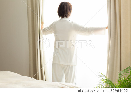 A middle-aged woman in pajamas opening the curtains in her bedroom in the morning 122961402