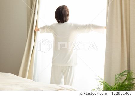 A middle-aged woman in pajamas opening the curtains in her bedroom in the morning 122961403