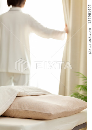 A middle-aged woman in pajamas opening the curtains in her bedroom in the morning A middle-aged woman in pajamas opening the curtains in her bedroom in the morning 122961405