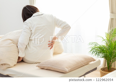 A middle-aged woman in white pajamas suffering from back pain in her bedroom in the morning A middle-aged woman in white pajamas suffering from back pain in her bedroom in the morning 122962054