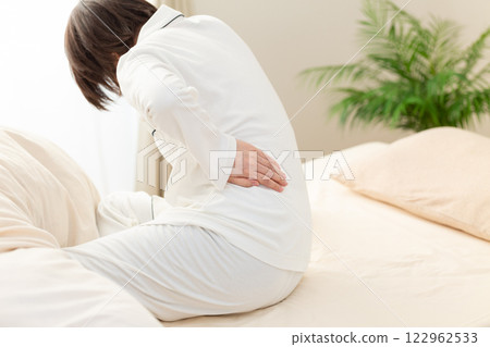 一名身穿白色睡衣的中年婦女早上在臥室裡感到背痛 一名身穿白色睡衣的中年婦女早上在臥室裡感到背痛 122962533