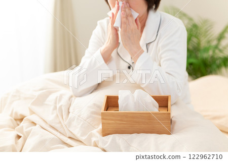 A middle-aged woman in white pajamas blowing her nose with a tissue after waking up 122962710