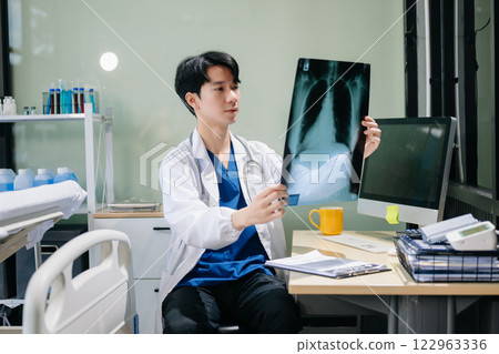 Confident young male doctor in white medical uniform sit at desk working on computer. 122963336