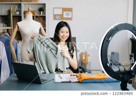 Asian lady blogger showing clothes in front of the camera to recording vlog video live streaming at her shop.  122963460