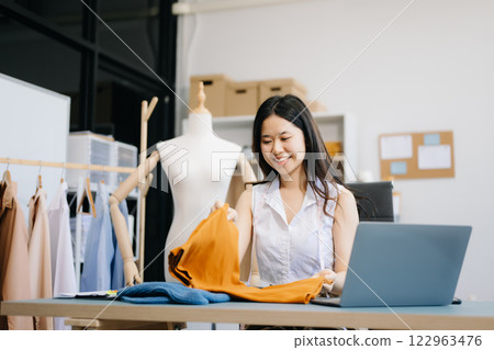 Asian young woman on desk in office of fashion designer and holds tablet, laptop and smartphone on desk Asian young woman on desk in office of fashion designer and holds tablet, laptop and smartphone on desk 122963476