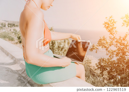 Woman Laptop Beach Working Remotely on Vacation - Beautiful young woman in a bathing suit using a laptop while sitting on a cliff overlooking the ocean. Woman Laptop Beach Working Remotely on Vacation - Beautiful young woman in a bathing suit using a laptop while sitting on a cliff overlooking the ocean. 122965519