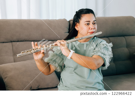 A female flutist plays a silver flute near a gray sofa in her living room, dressed in a casual green shirt. The serene setting emphasizes her musical talent and love for the arts. 122966422