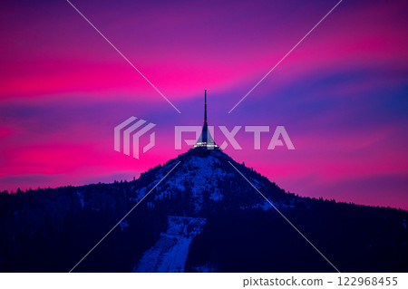 Jested Mountain stands majestically against a vibrant winter evening sky. The peak is illuminated as colorful clouds reflect shades of purple and blue, creating a serene atmosphere. Jested Mountain stands majestically against a vibrant winter evening sky. The peak is illuminated as colorful clouds reflect shades of purple and blue, creating a serene atmosphere. 122968455