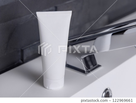 White cream tube near modern faucet on basin in dark grey bathroom close up, cosmetic mockup 122969661