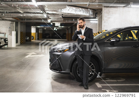Caucasian male business professional holding money and speaking on phone in underground parking garage. Dressed in formal suit, exhibiting confidence. 122969791