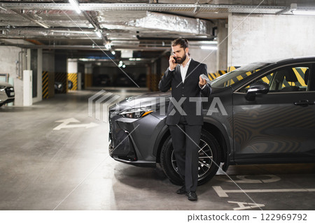 Caucasian male business professional holding money and speaking on phone in underground parking garage. Dressed in formal suit, exhibiting confidence. 122969792