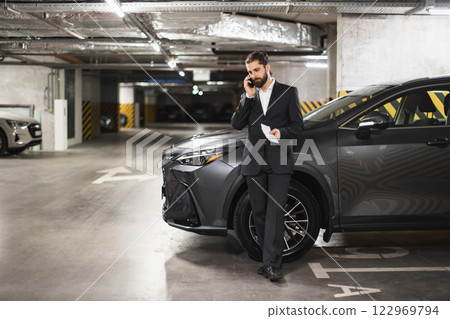 Young adult Caucasian male in formal attire talks on phone holding cash, standing by car in underground parking. Scene conveys business and prosperity. Young adult Caucasian male in formal attire talks on phone holding cash, standing by car in underground parking. Scene conveys business and prosperity. 122969794
