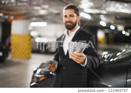 Young Caucasian male businessman counting cash in underground parking lot. Image conveys wealth, secrecy, and business themes in urban environment. Young Caucasian male businessman counting cash in underground parking lot. Image conveys wealth, secrecy, and business themes in urban environment. 122969797