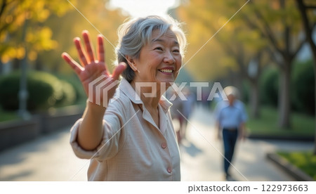Elderly woman waving in a sunlit park during autumn, surrounded by colorful trees and other visitors 122973663