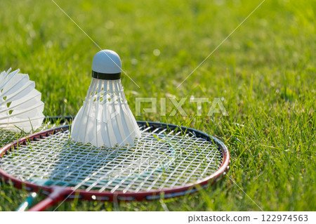 White badminton shuttlecock and badminton rackets on green grass in sunny shadow. Summer. Leisure games. Outdoors. Close up. 122974563