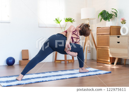 Senior Asian woman practicing Parivrtta Parsvakonasana Revolved Side Angle Pose on a yoga mat in a cozy home setting Senior Asian woman practicing Parivrtta Parsvakonasana Revolved Side Angle Pose on a yoga mat in a cozy home setting 122975611