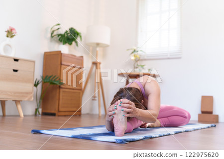 Senior Asian woman performing Janu Sirsasana Head-to-Knee Forward Bend Pose on a yoga mat in a peaceful living space Senior Asian woman performing Janu Sirsasana Head-to-Knee Forward Bend Pose on a yoga mat in a peaceful living space 122975620