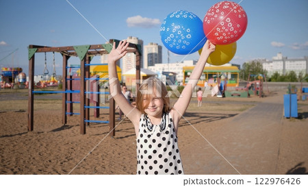 A joyful child wearing a polka dot dress holds a bunch of colorful balloons at a vibrant playground filled with fun 122976426