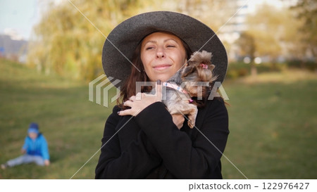 A joyful woman is happily holding her small dog in a beautiful park while enjoying a lovely sunny day A joyful woman is happily holding her small dog in a beautiful park while enjoying a lovely sunny day 122976427