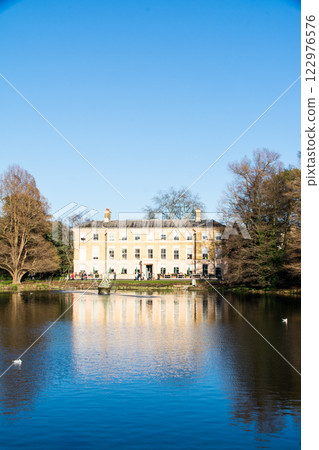 A historic building made of light brown bricks standing right next to a pond lit by bright sunlight. Kewgarden, a suburb of London A historic building made of light brown bricks standing right next to a pond lit by bright sunlight. Kewgarden, a suburb of London 122976576